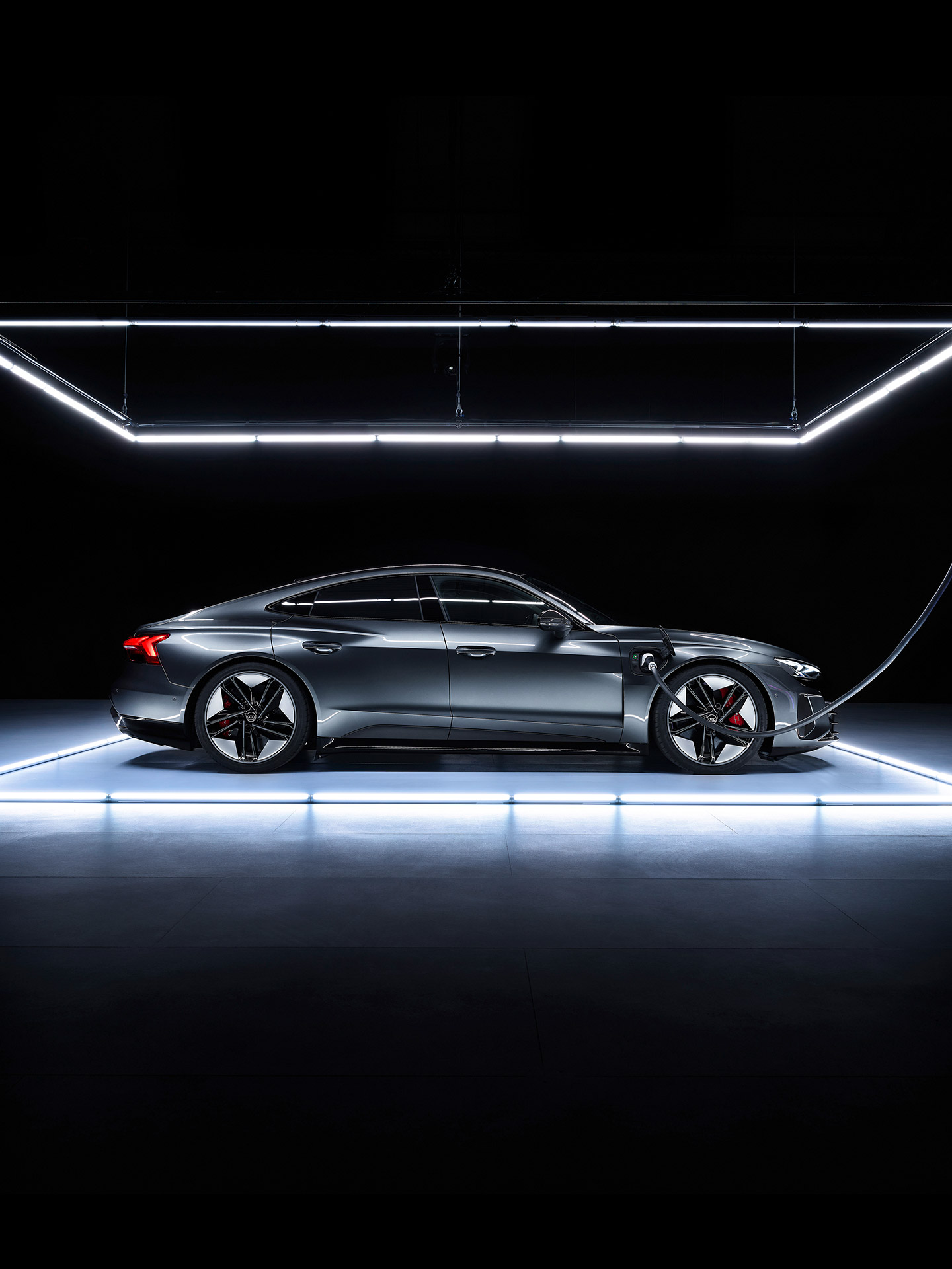 Shifting image of a gray Audi e-tron GT parked in front of a daytime city skline and on an illuminated platform in a dark room. 