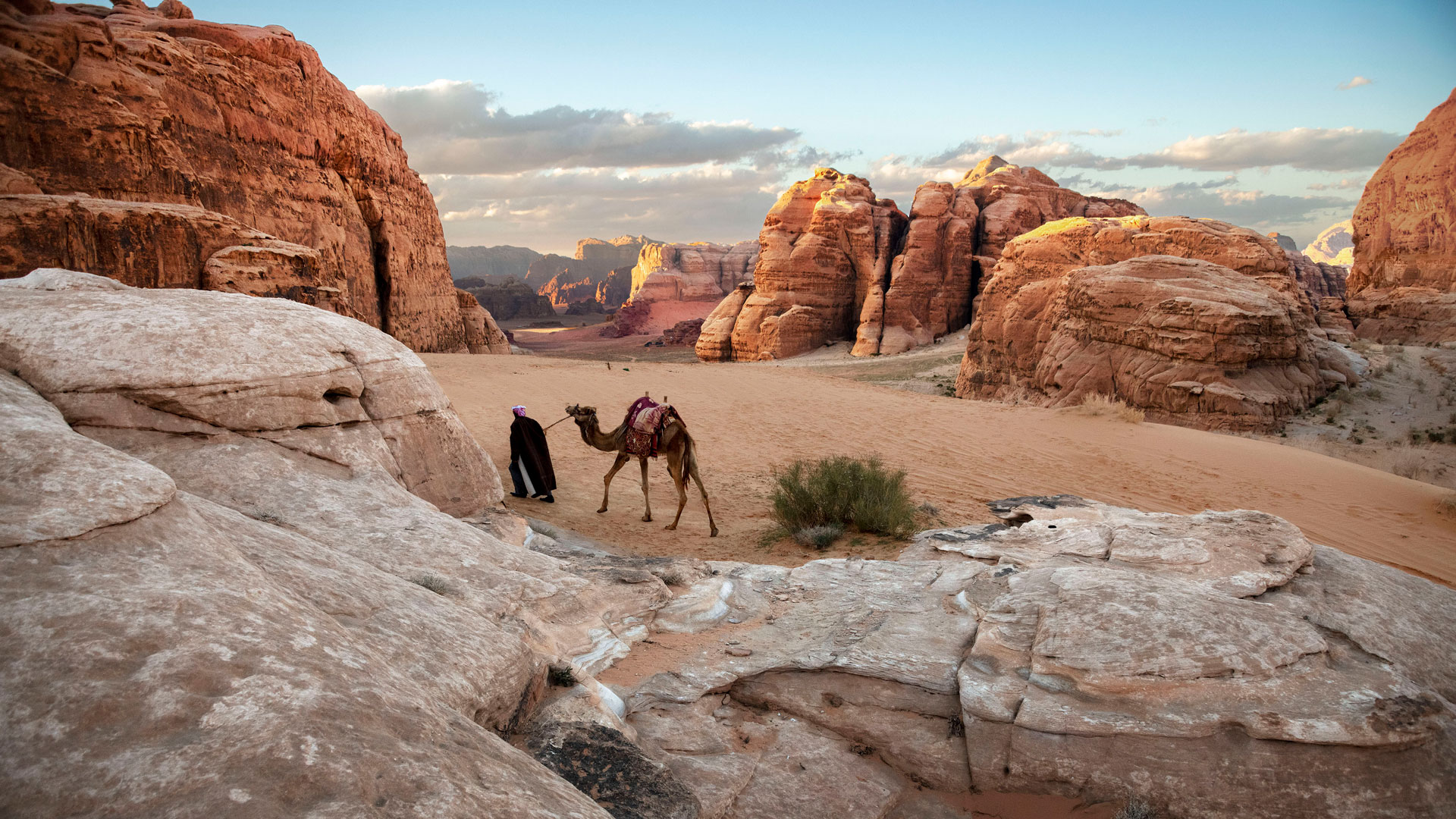 A desert scene featuring a man leading a camel through a red, rocky landscape.